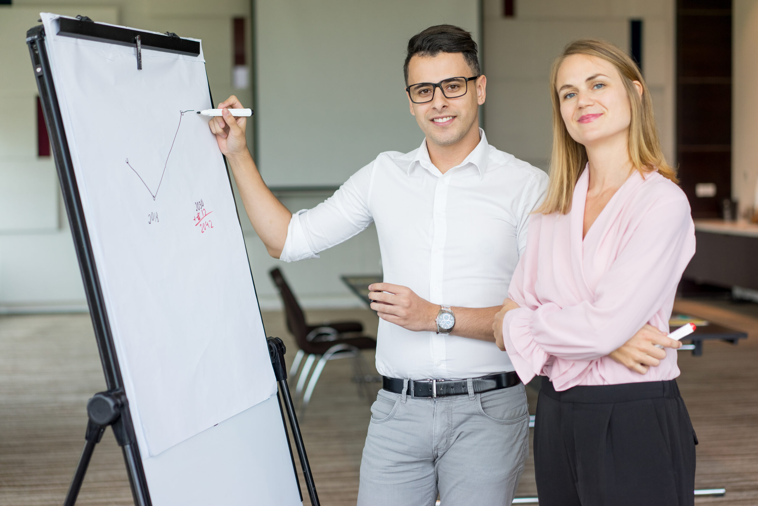 Portrait of smiling business colleagues drawing plan on flipchart. Young Caucasian businessman wearing glasses and businesswoman smiling at camera in boardroom. Teamwork and briefing concept